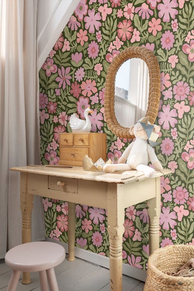 Blumige Vliestapete in Rosa und Lila auf grünem Grund in einem Kinderzimmer mit Holzschreibtisch und Rattanspiegel.