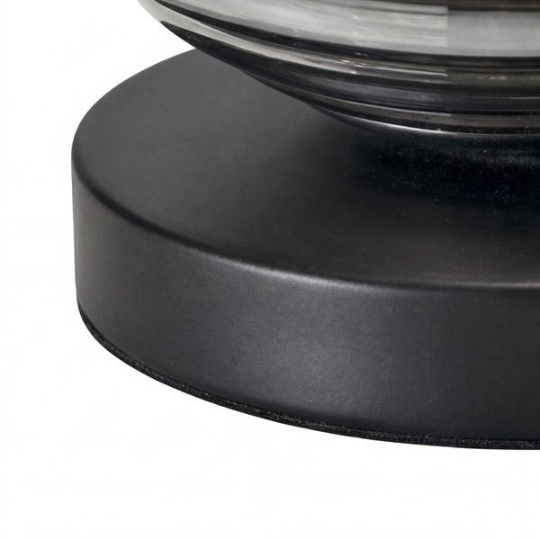 Detail eines runden, schwarzen Lampenfußes aus mattem Metall mit einem Glaselement.