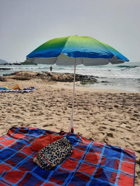 Sonnenschirm mit blau-grünem Farbverlauf auf einem Sandstrand mit Picknickdecke im Vordergrund.