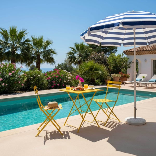 Helle Gartenszene mit Außenpool, gelber Bistro-Garnitur aus Metall und gestreiftem Sonnenschirm zwischen Palmen und blühenden Sträuchern.