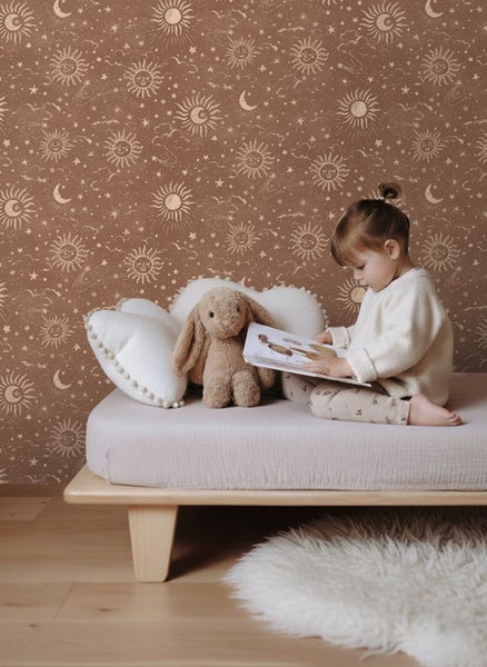 Kindertapete mit Sonnen, Mond und Sternen in Rostbraun, Kind liest Buch auf Bett mit Kuscheltier Hase und weißen herzförmigen Kissen.