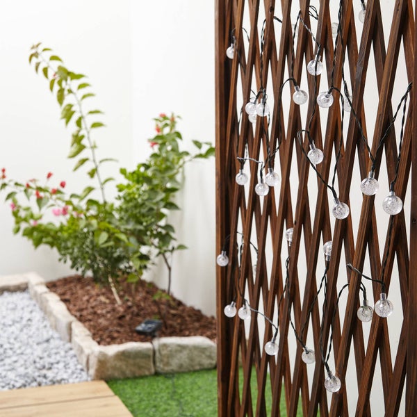 Solar-LED-Lichterkette mit klaren Kugeln in Blasenoptik an einem braunen Rankgitter aus Holz in einem hellen Garten mit Pflanzen.