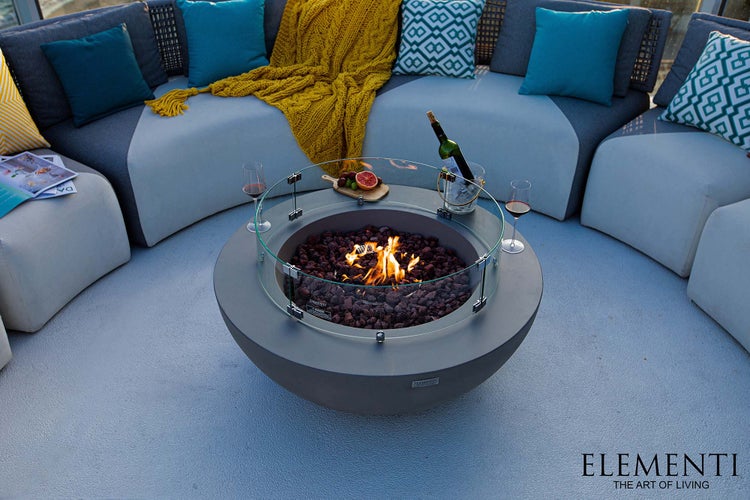 Moderne runde Gasfeuerstelle aus Beton mit Glasschutz und Lavasteinen in Lounge-Ecke mit Sofa, Kissen und Decke. Elementi Logo.