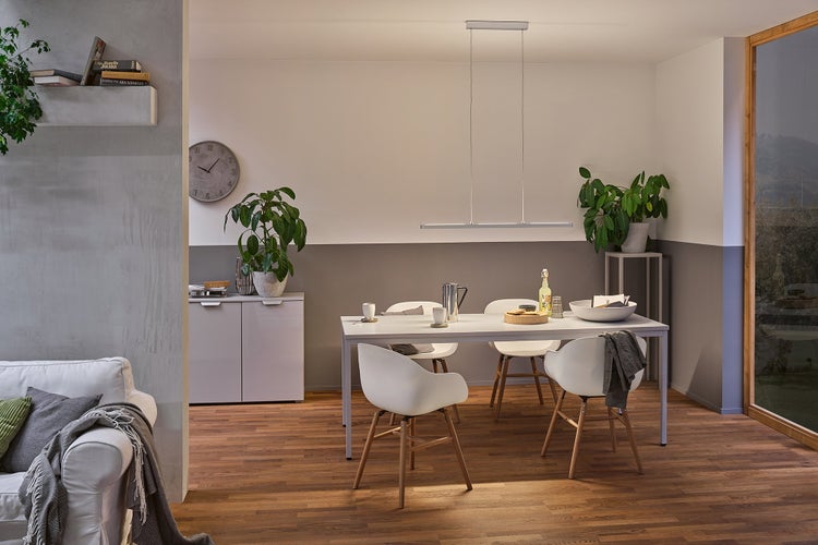 Heller Essbereich mit weißem Tisch, Designerstühlen, einer LED-Pendelleuchte, Zimmerpflanzen und Boden in Holzoptik.
