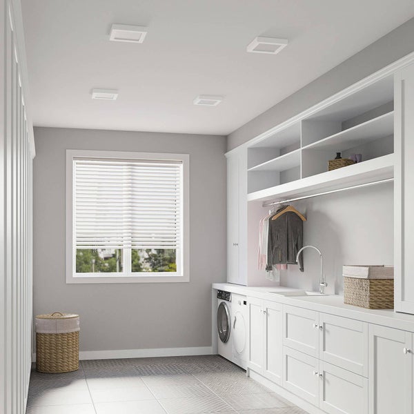 Moderner, heller Hauswirtschaftsraum mit vier quadratischen LED-Deckenpanelen, weißen Einbauschränken, einer Waschmaschine und Fenster mit Jalousien.