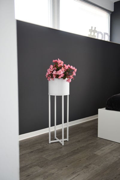 Moderner hoher weißer Pflanzenständer aus Metall mit weißem Übertopf und rosa Begonien in einem Raum mit Boden in Holzoptik.