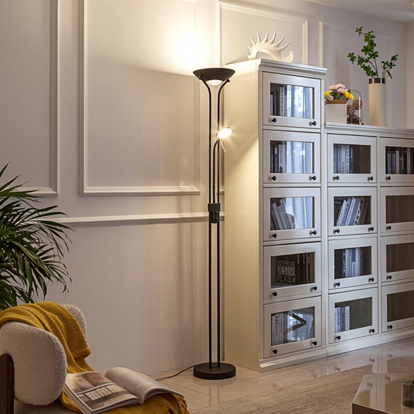 Schwarze Deckenfluter Stehleuchte aus Metall mit integrierter Leseleuchte in einem hellen Wohnzimmer neben einem weißen Regal.