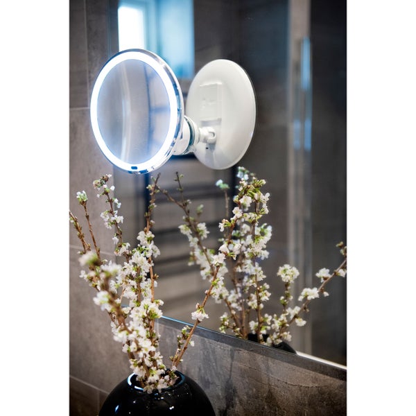 Runder LED Kosmetikspiegel mit Wandhalterung vor einem Badezimmerspiegel mit blühenden Zweigen im Vordergrund.
