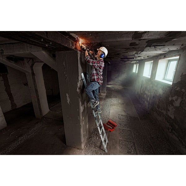 Handwerker arbeitet mit einem Winkelschleifer an einer Betonsäule auf einer Aluminiumleiter in einem unfertigen Kellerraum.