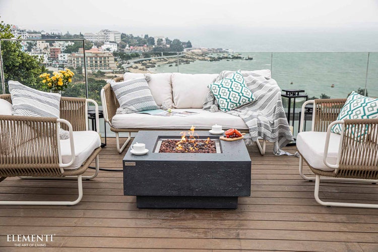 Moderner Gas-Feuertisch aus Beton auf einer Holzterrasse mit Gartenmöbeln, Kissen und Blick auf das Meer.