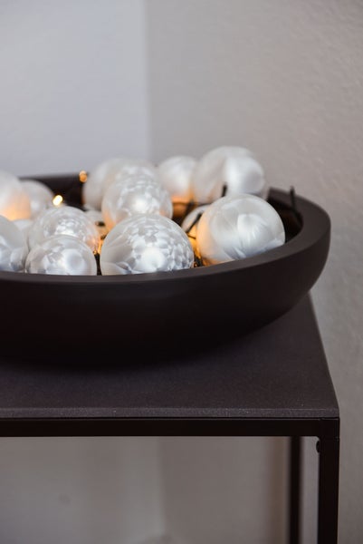 Dunkle Dekoschale gefüllt mit strukturierten weißen Weihnachtskugeln und einer warmweißen Lichterkette auf einem Beistelltisch.