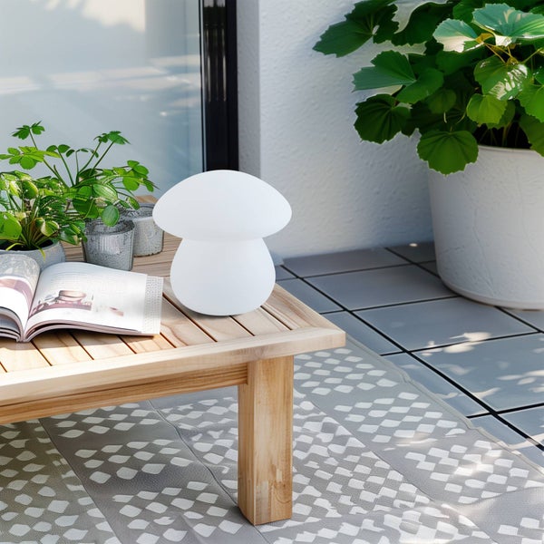 Pilzförmige Tischleuchte auf einem Holztisch im Außenbereich, neben einer Zeitschrift und Pflanzen.