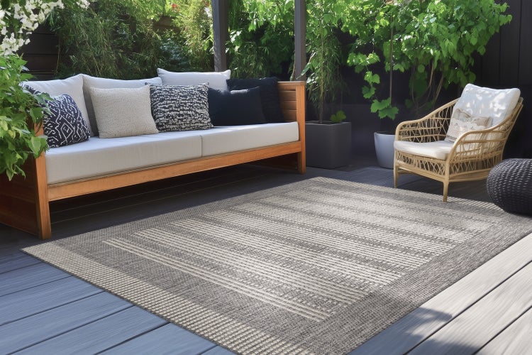 Outdoor-Teppich mit Streifenmuster auf einer Holzterrasse vor einem Garten-Sofa und einem Korbsessel.
