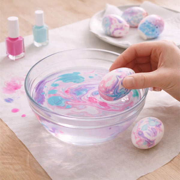 Hand taucht ein Ei in eine Schüssel mit Wasser und Nagellack zum Marmorieren. Fertige marmorierte Ostereier und Nagellackflaschen liegen auf dem Tisch.