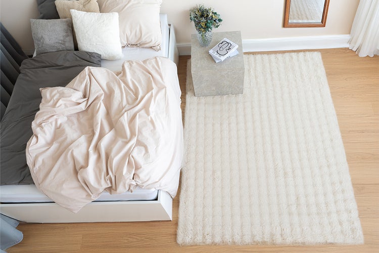 Ein moderner Schlafbereich mit einem weichen Hochflor Teppich, einem Bett mit mehrfarbiger Bettwäsche, einem Nachttisch und Holzboden.
