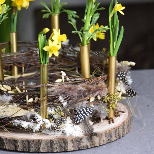 Frühlingsgesteck auf Holzscheibe mit Zweigenest, goldenen Röhrenvasen, Osterglocken, Federn und Trockenblumen.