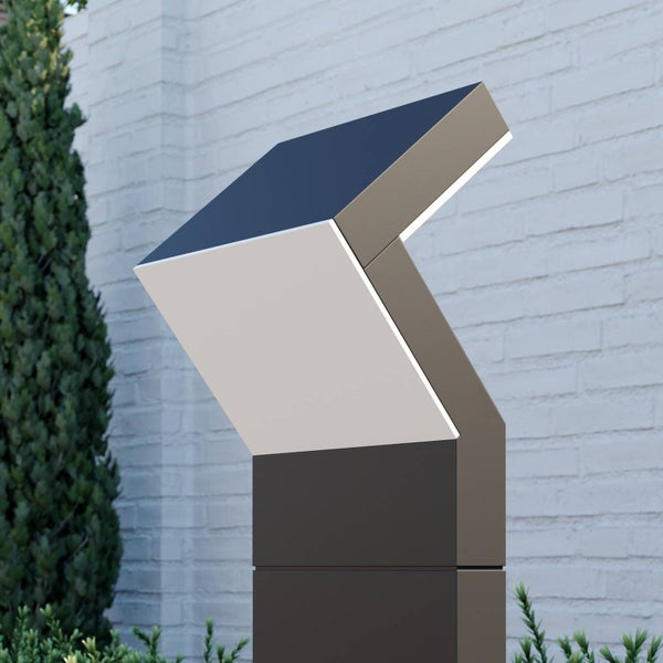 Moderne geometrische Solar-Wegeleuchte für den Außenbereich mit weißer Lichtfläche vor einer hellen Ziegelwand und grünen Pflanzen.