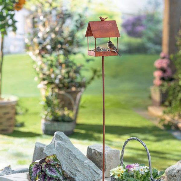Vogelhaus aus Metall in Rostoptik auf einem Stab in einem sonnigen Garten mit einem Vogel auf der Futterfläche.