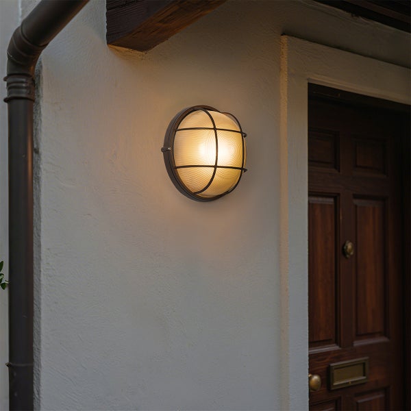 Runde Außenwandleuchte mit Metallgitter und Glasabdeckung, montiert an einer Hauswand neben einer Holztür.
