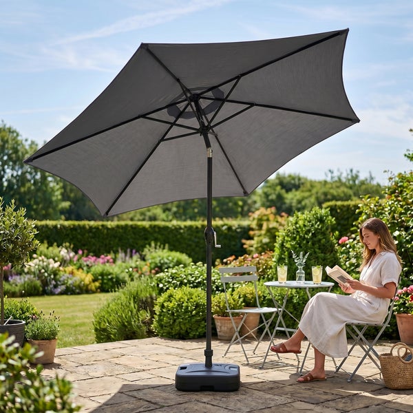 Grauer Garten-Sonnenschirm mit Kurbel und Schirmständer auf einer Terrasse in einem blühenden Garten mit Frau beim Lesen.