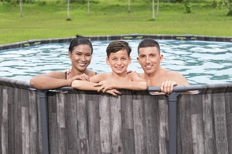 Dreiköpfige Familie in einem runden Aufstellpool mit grauer Holzoptik in einem Garten.
