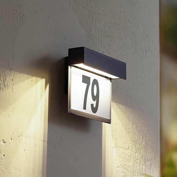 Beleuchtete Hausnummernleuchte mit dunklem Rahmen und der Zahl 79, montiert an einer Wand.