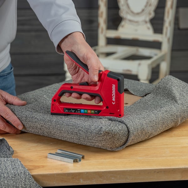 Handtacker mit Arrow Logo befestigt Polsterstoff an einem Sitzkissen. Die Szene auf dem Holztisch ist hell und organisiert.