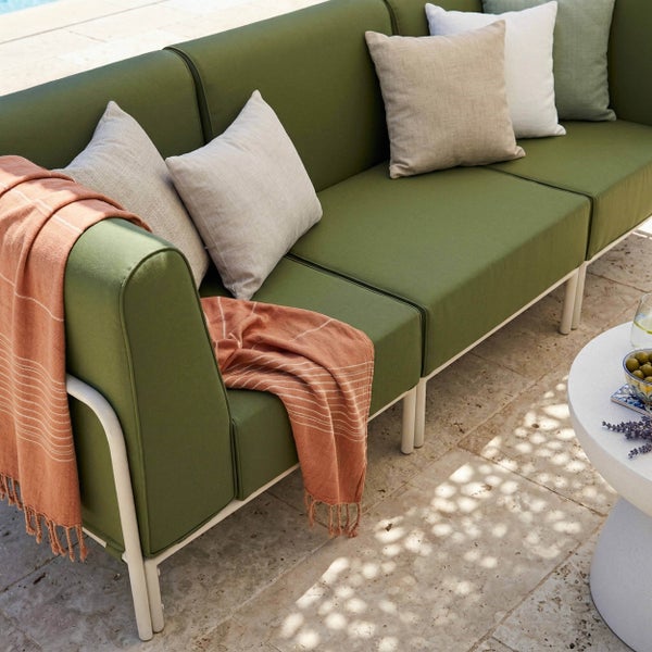 Modernes grünes Outdoor-Sofa mit weißem Metallgestell, dekoriert mit verschiedenen Kissen und einer orangefarbenen Decke auf einer Steinterrasse.