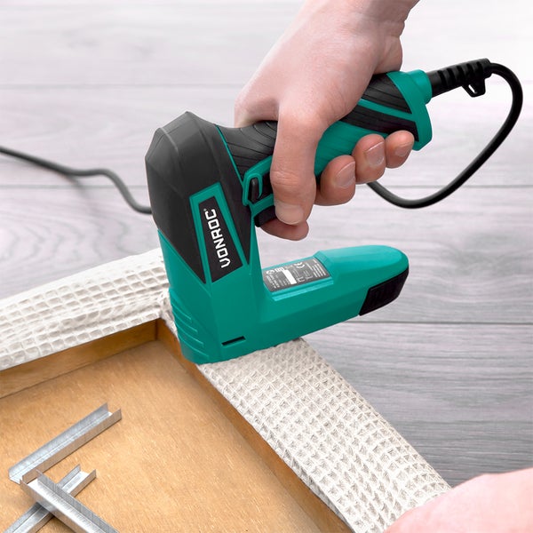 Eine Hand bedient einen elektrischen Tacker zum Befestigen von Stoff an einem Holzrahmen, wobei sich Tackernadeln in der Nähe befinden.