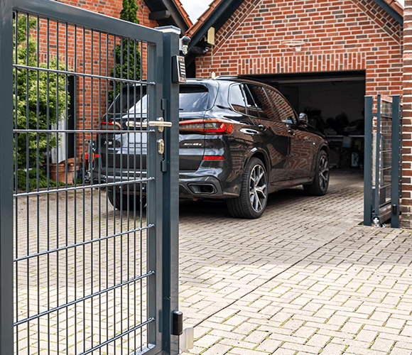 Geöffnetes Doppelstabmatten-Tor in Anthrazit vor einer gepflasterten Einfahrt mit Auto und einer hellen Backsteingarage.