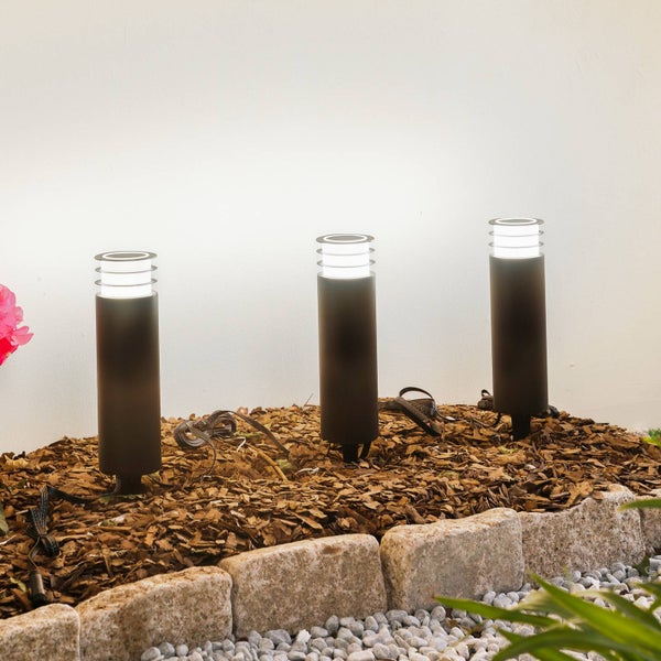 Drei schwarze zylindrische Außenleuchten in einem Beet mit Rindenmulch und Steinkante, dargestellt in warmweißem, neutralweißem und kaltweißem Licht.