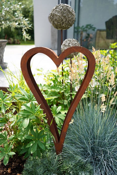 Gartenstecker in Herzform aus Metall mit Edelrost Patina in einem Gartenbeet zwischen Pflanzen.