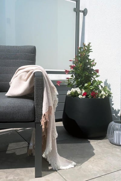 Grauer Gartensessel mit hellbeige Wolldecke, großer schwarzer Pflanzkübel mit Blumen und eine Laterne auf einer gefliesten Terrasse.