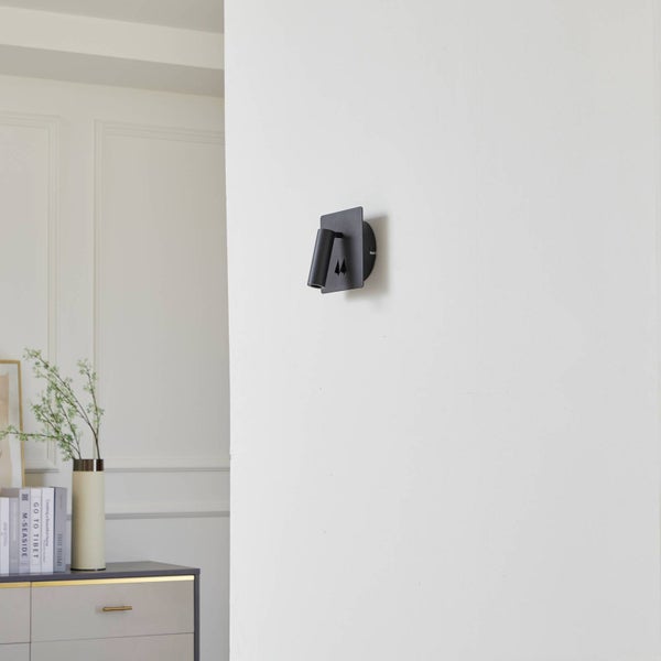 Moderne schwarze Wandleuchte mit verstellbarem Spot an einer weißen Wand in einem hellen Wohnraum.