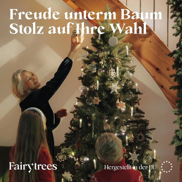 Eine Frau und zwei Kinder schmücken einen Fairy Trees Weihnachtsbaum mit Dekorationen in einem Wohnzimmer neben einer Holztreppe.