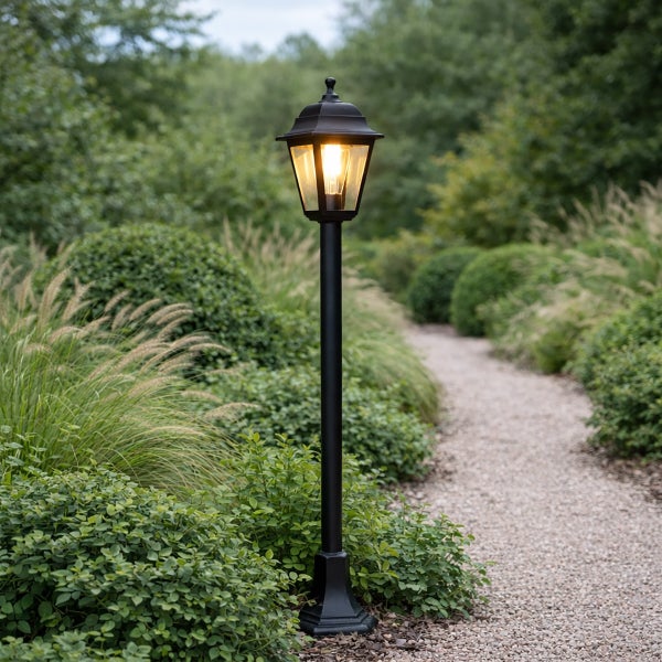 Klassische schwarze Gartenlaterne aus Metall auf einem Pfosten neben einem organisierten Kiesweg, umgeben von Pflanzen und Ziergräsern.
