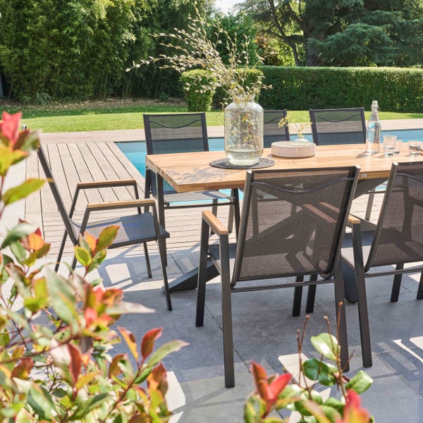 Hell gestalteter Außenbereich mit Holztisch und dunkelgrauen Stapelstühlen aus Metall und Textilene auf einer Terrasse am Pool im grünen Garten.