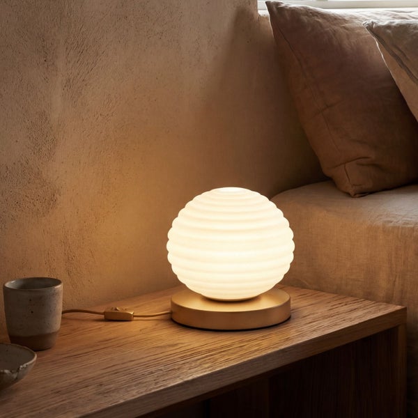 Tischleuchte mit weißem, geripptem Kugelschirm und goldfarbenem Metallsockel auf einem Holztisch, warmes Licht ausstrahlend.