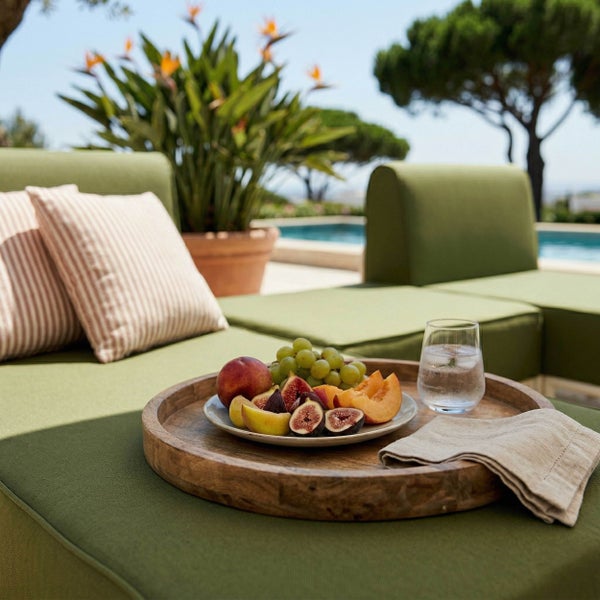 Holztablett mit Obst und Wasserglas auf grünem Garten-Lounge-Sofa mit gestreiften Kissen im Außenbereich am Pool.