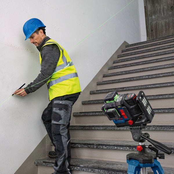 Mann mit Laser-Wasserwaage und Sicherheitsausrüstung beim Markieren an einer Wand auf einer Treppe.