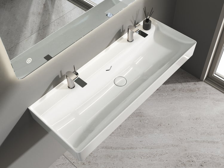 Modernes, helles Badezimmer mit breitem Doppelwaschbecken aus weißer Keramik, zwei verchromten Armaturen und einem großen, hinterleuchteten Spiegel.