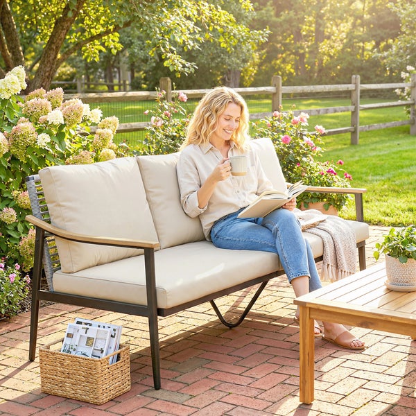 Frau liest auf einem modernen 2-Sitzer Gartensofa mit beigen Polstern und Holzarmlehnen auf einer hellen, gepflasterten Terrasse.