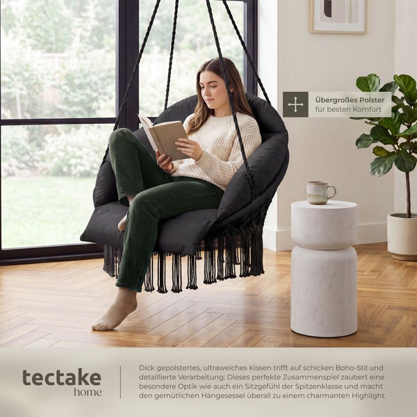 Frau liest in einem Hängesessel im Boho-Stil mit dickem Kissen und Fransen. Helles Wohnzimmer mit Holzfußboden. tectake home Logo.