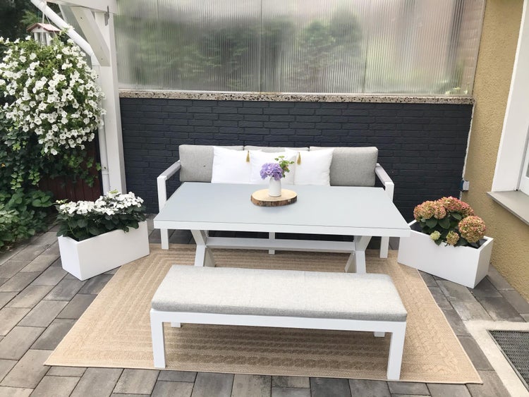 Modernes Gartenmöbel-Set mit weißem Esstisch, Sofa und Sitzbank mit grauen Polstern auf einem beigefarbenen Teppich auf einer Terrasse.