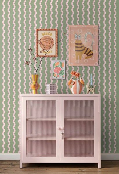 Vliestapete mit grün-rosa Wellenmuster in einem hellen Zimmer mit rosa Metallschrank, Wandbildern und farbenfroher Dekoration im Retro-Stil.