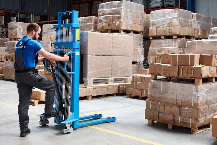 Ein Mitarbeiter bedient einen blauen manuellen Hochhubwagen in einem organisierten Lager, um eine Palette mit Kartons anzuheben.