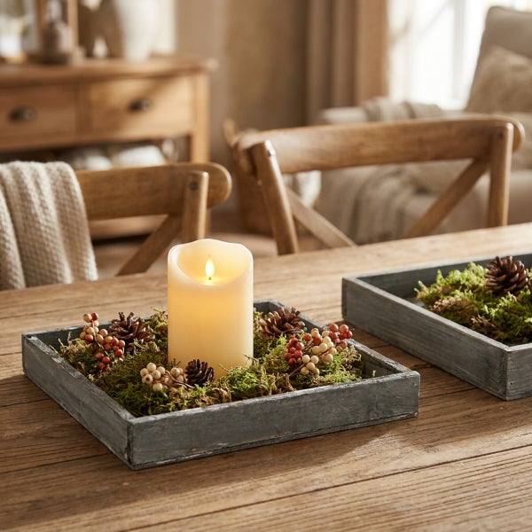 LED Kerze in einem quadratischen Holztablett dekoriert mit Moos, Tannenzapfen und Beeren auf einem rustikalen Holztisch.
