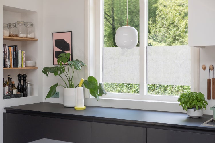 Helle, organisierte Küche mit Fenster, Sichtschutz, Zimmerpflanzen und eingebauten Regalen auf einer dunklen Arbeitsplatte.