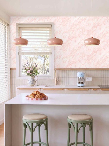 Helle, moderne Küche mit einer marmorierten Tapete in Rosa, drei Pendelleuchten, einer Kücheninsel mit Hockern und einer Kaffeemaschine.