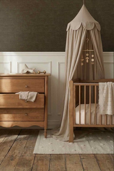 Kinderzimmer mit dunkler Strukturtapete und weißer Wandvertäfelung. Mit Holzkommode, Babybett mit Stoffbaldachin und Holzdielenboden.